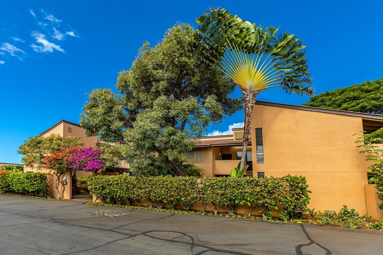 A building with a beige exterior, surrounded by lush greenery and a clear blue sky.
