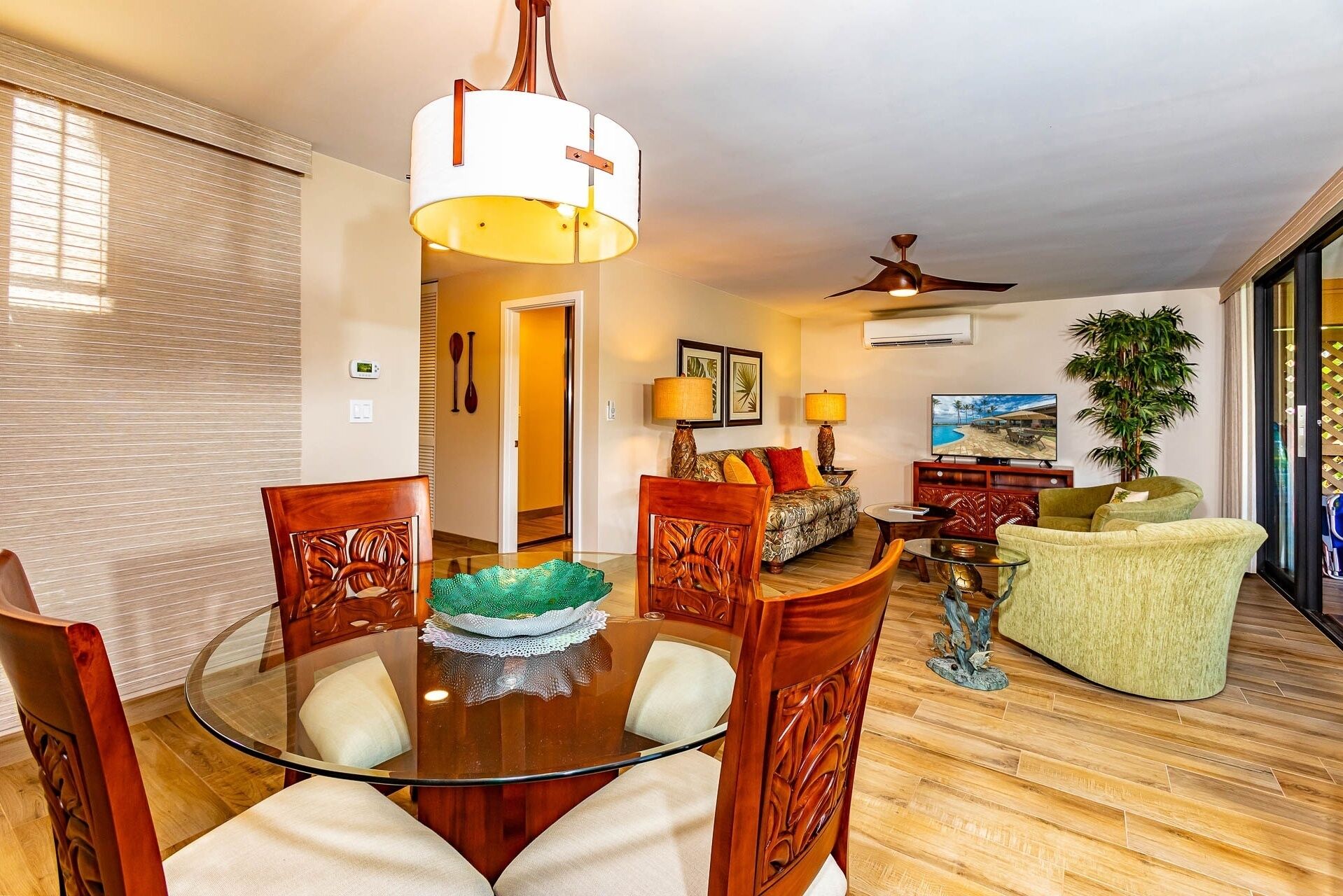 A well-lit living room with a dining area, a ceiling fan, and a TV displaying a tropical scene.