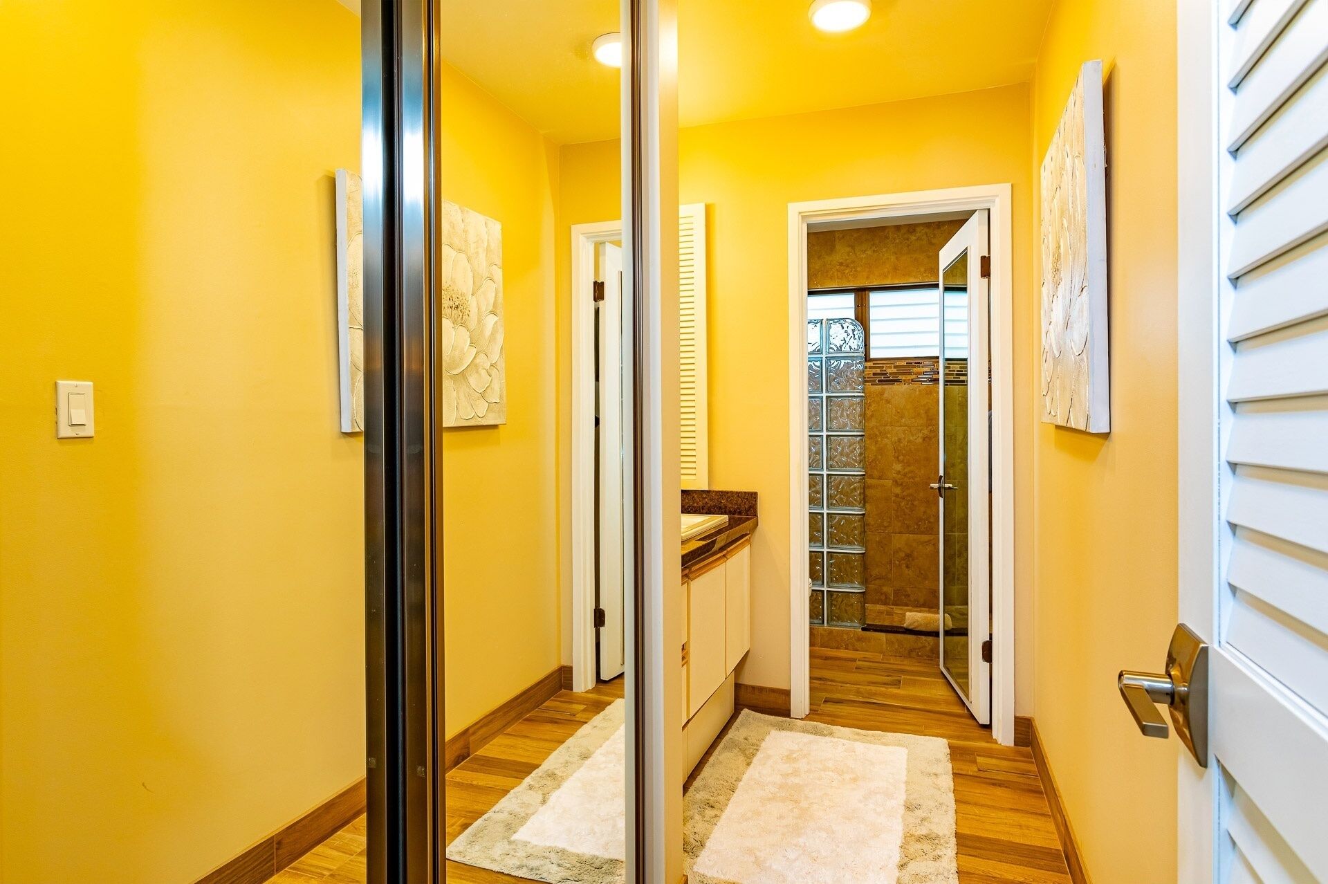 Un pasillo con paredes amarillas, suelo de madera y una puerta de cristal que da a un baño.