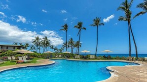 Una piscina con sillas de descanso y sombrillas, rodeada de palmeras y un cielo despejado.