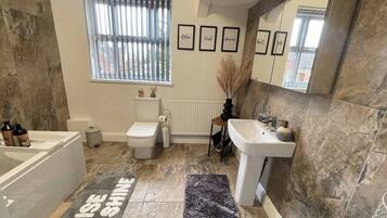 A bathroom with a bathtub, toilet, sink, and a window with blinds. There are framed pictures on the wall and a rug with text.