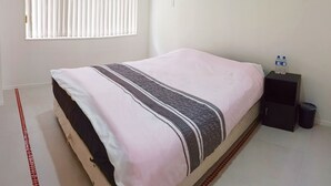 Un dormitorio con cama, ventana con persianas, mesita de noche con botellas de agua y un cesto de basura.