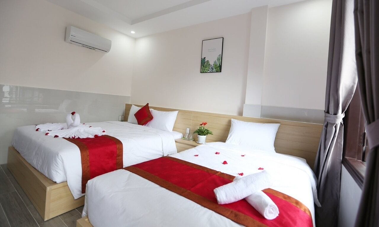 A hotel room with two beds, each with white bedding and red accents, a small plant on the nightstand, and a framed picture on the wall.