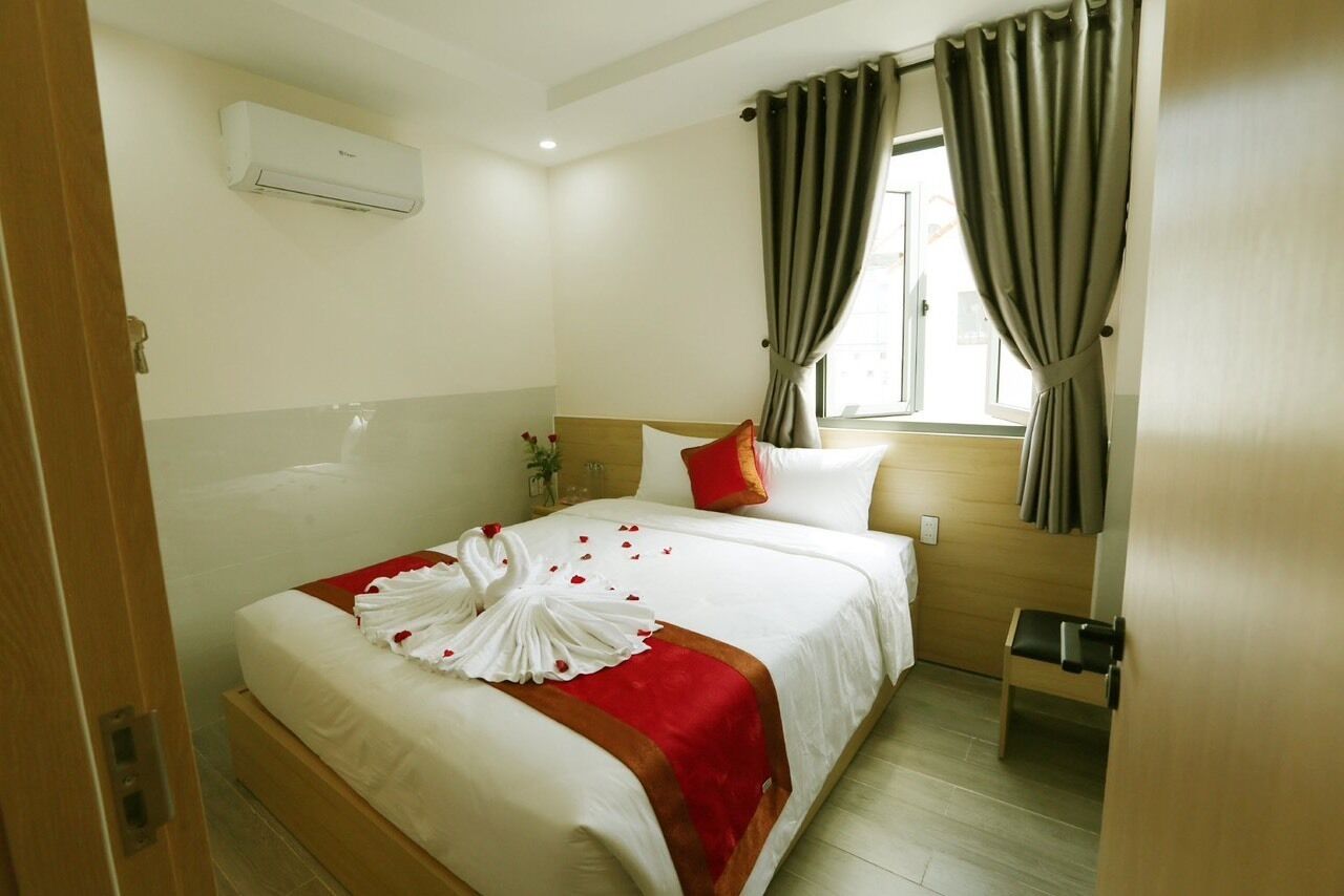 A hotel room with a neatly made bed, a window with curtains, an air conditioning unit, and a bedside table.