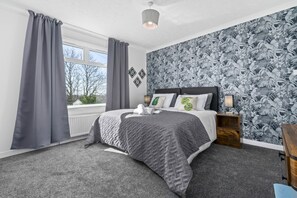 Um quarto com cama, cortinas cinzas, uma janela, parede com padrão de folhas e uma mesa de cabeceira com abajur.