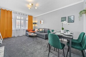 A modern living room with a grey sofa, orange curtains, and a dining area with green chairs and a table set for four.