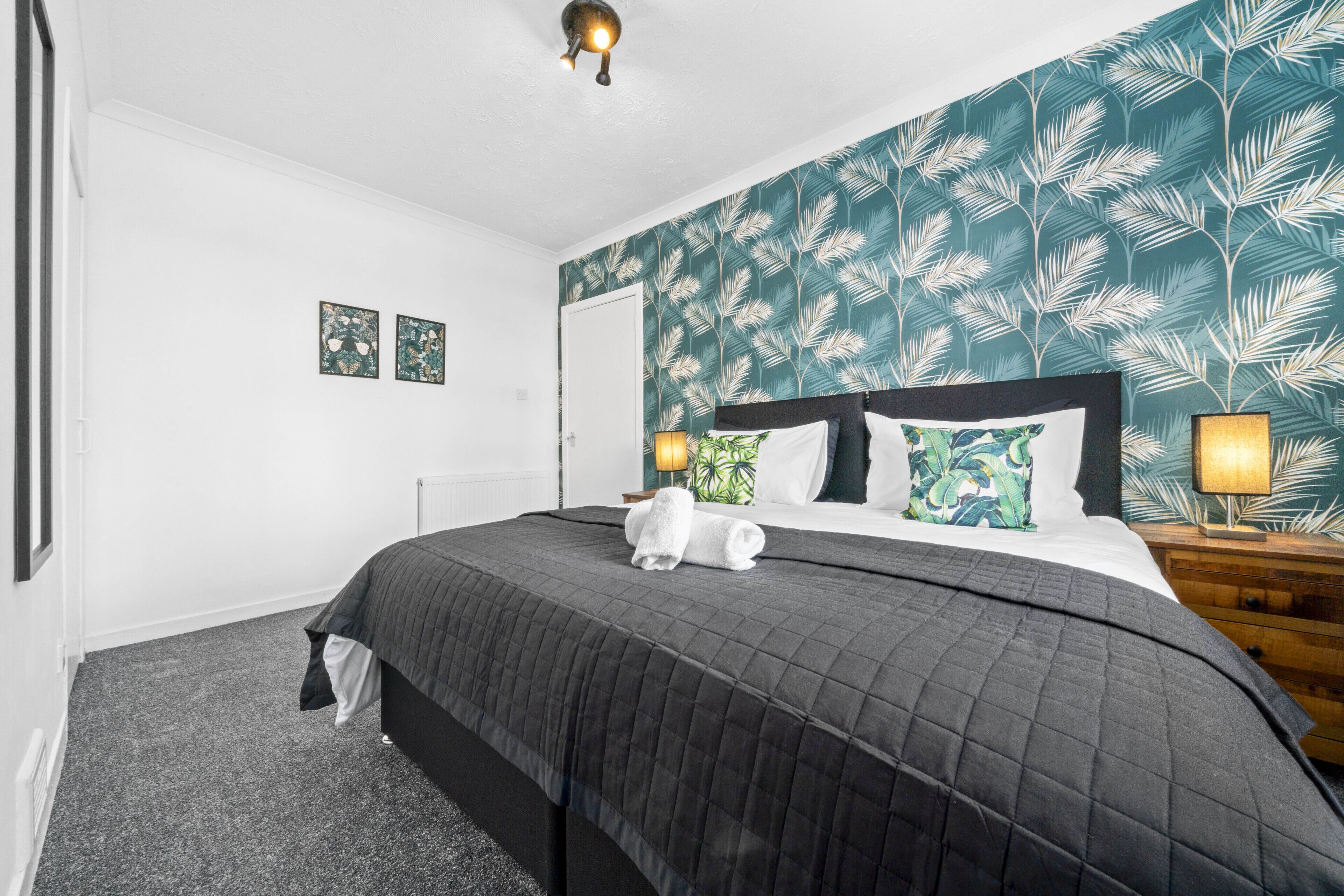 A neatly made bed with a dark quilt, two pillows, and a rolled-up towel. There are two bedside lamps on wooden nightstands. The room has a white door and a wall with a leafy pattern.