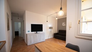 A modern living room with a wooden dining table, a flat-screen TV on a white cabinet, a dark sofa, and a floor lamp. - Appartamento Golf Resort (Manerba del Garda)