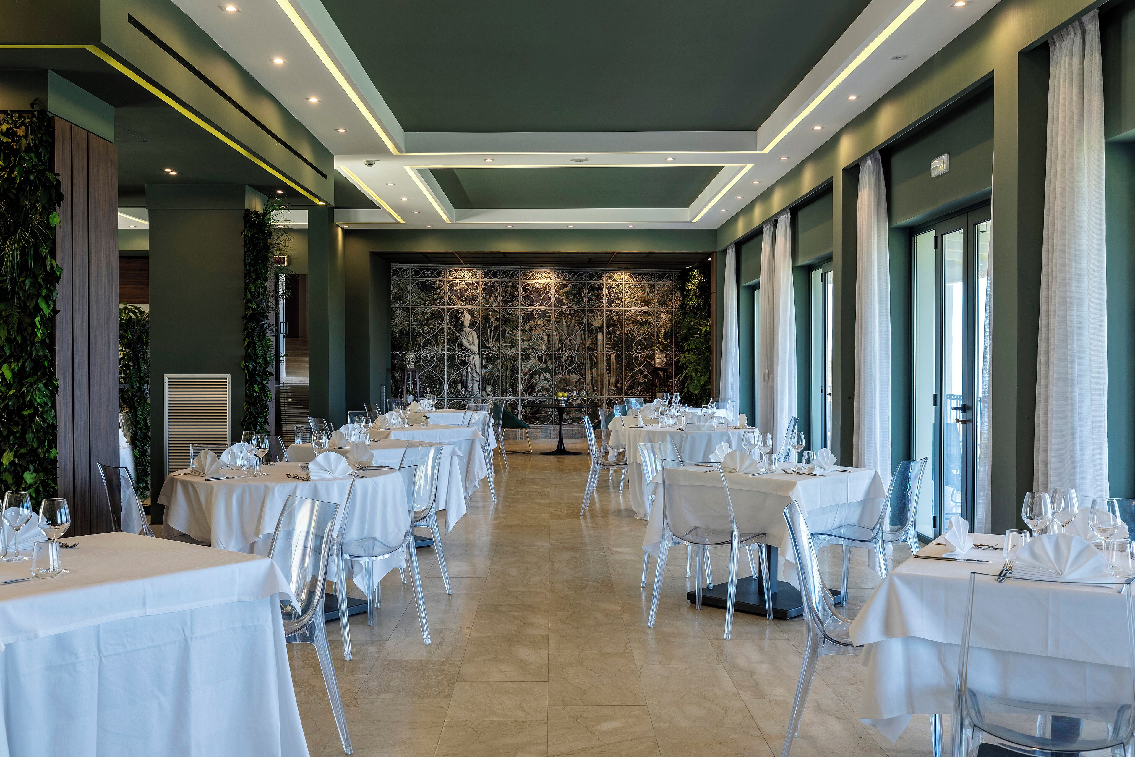 A restaurant with multiple tables set for dining, clear chairs, and a decorative wall in the background.