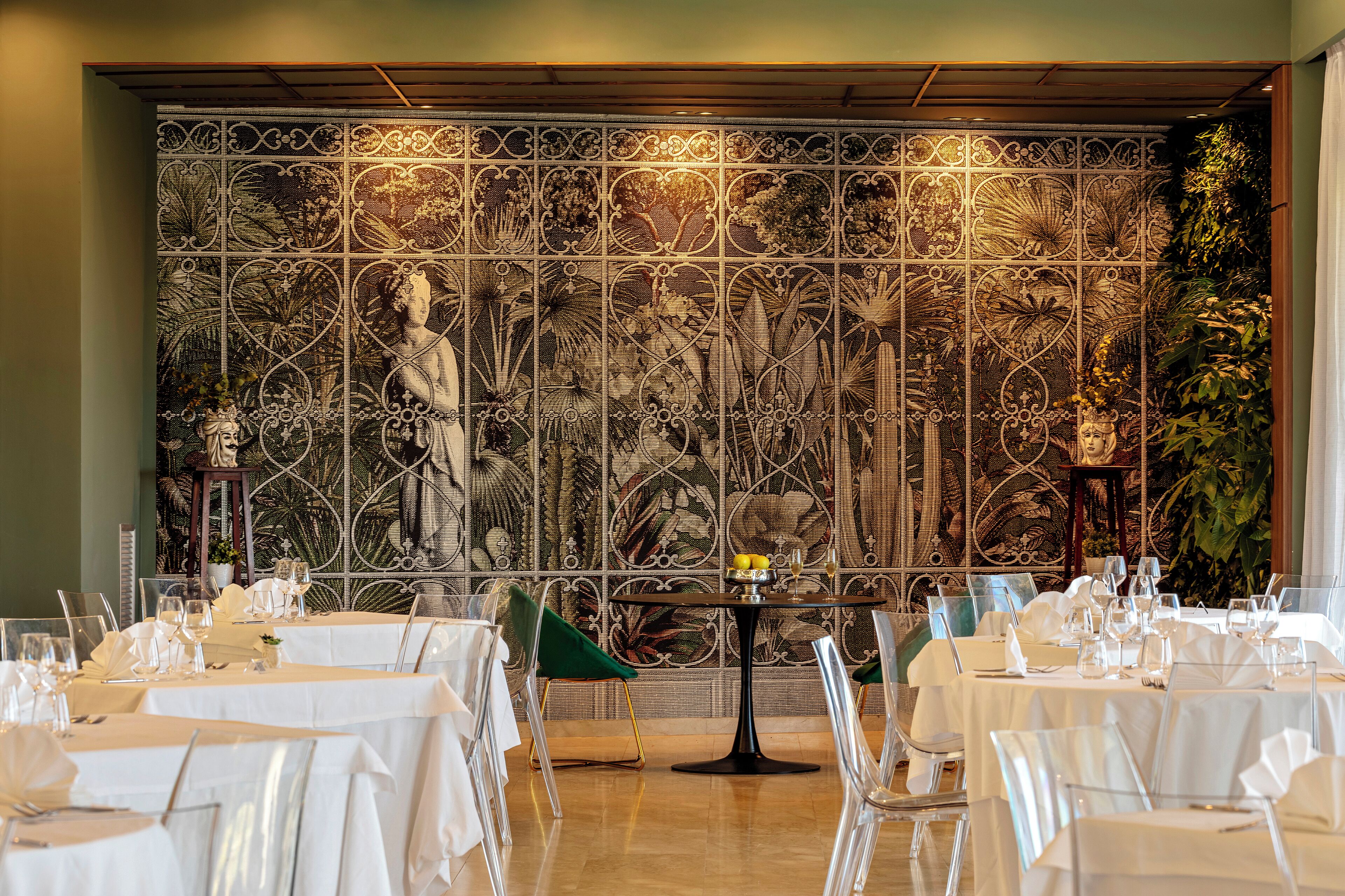 A dining area with tables set for a meal, a decorative wall with a floral pattern, and a plant on the right.