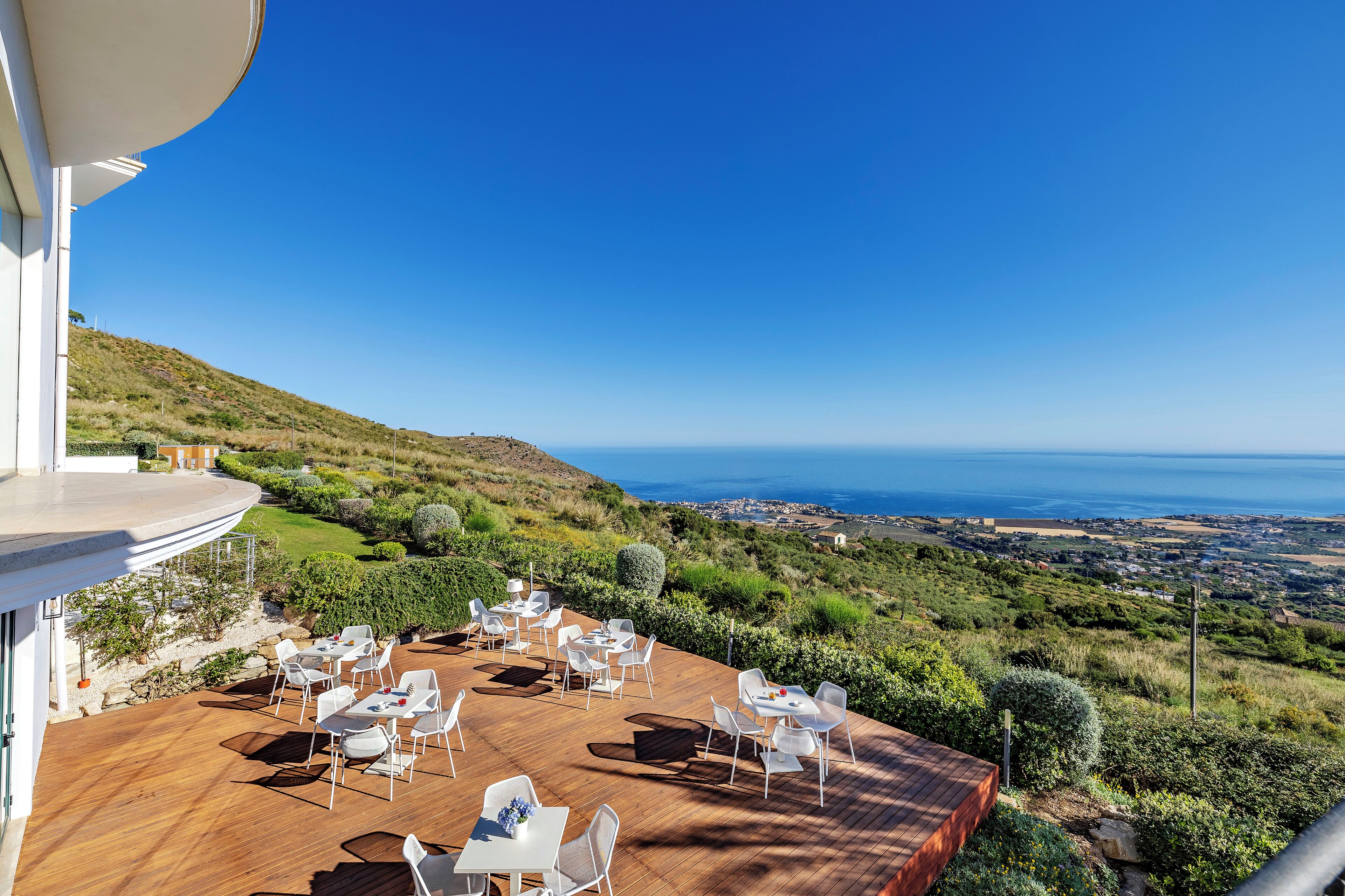 Una terrazza all'aperto con tavoli e sedie bianche, con vista su un paesaggio costiero.
