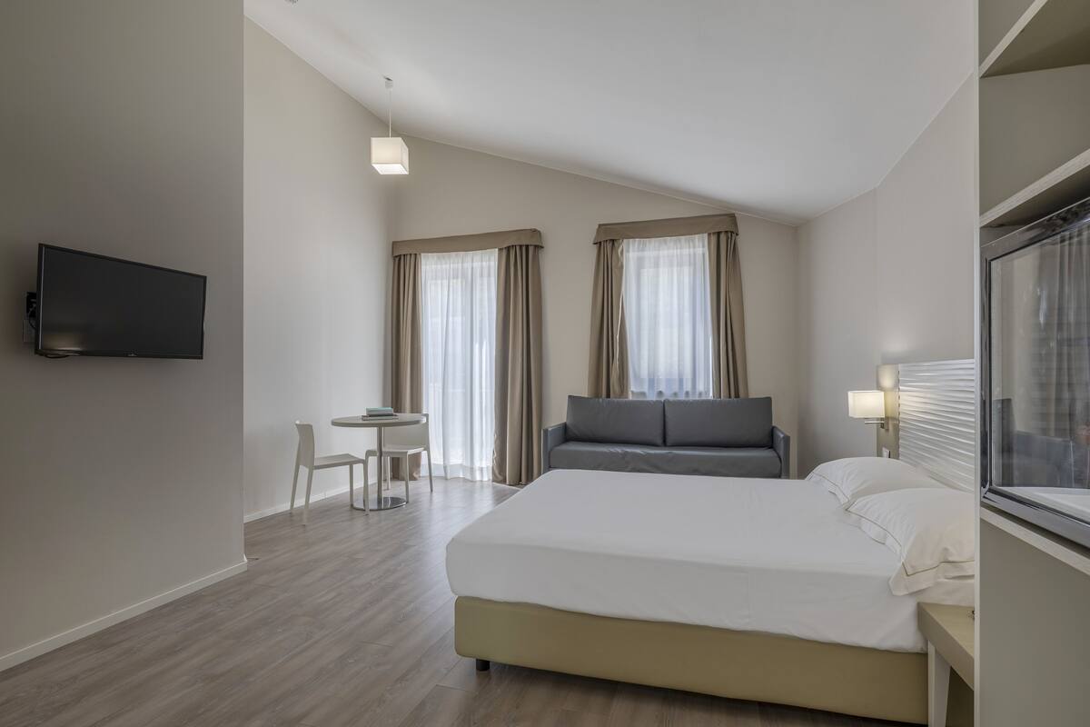 Habitación de hotel con cama, escritorio, silla, televisor y ventana con cortinas.