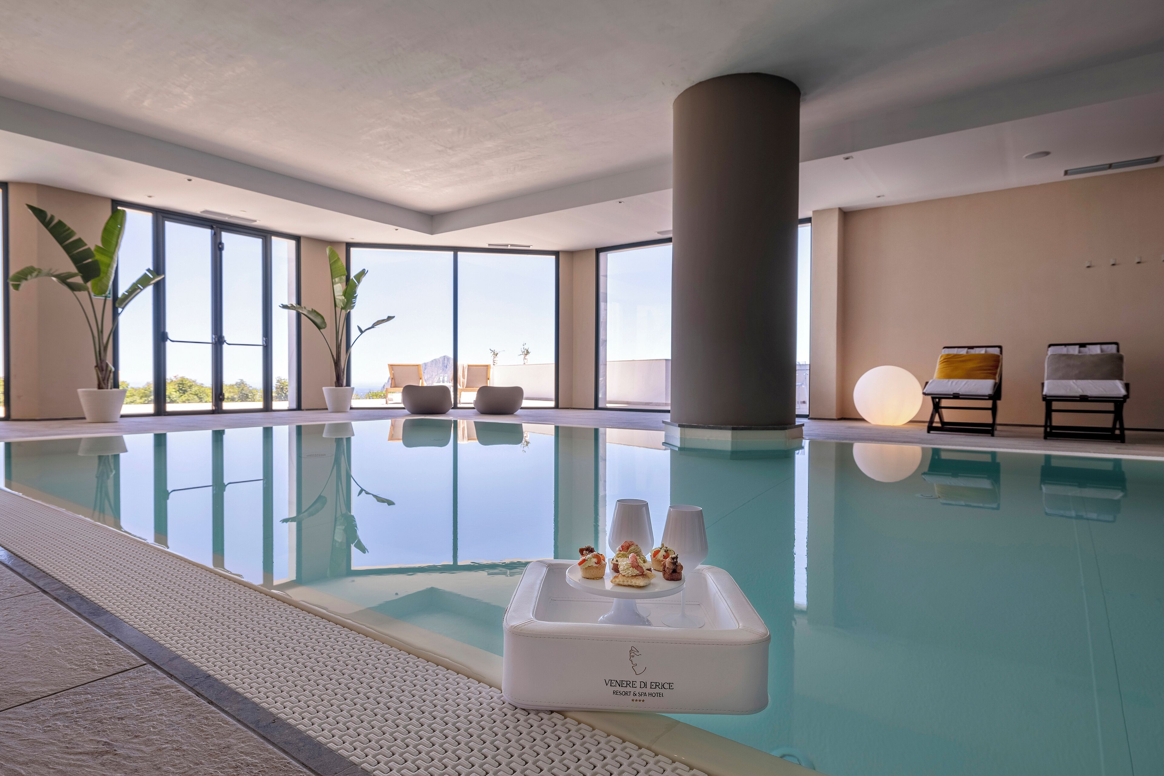 An indoor swimming pool with clear glass walls, a white lounge area, and a table with snacks.
