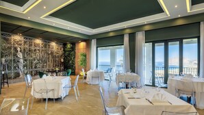 A restaurant with clear glass chairs and tables, a mural on the wall, and a view of the sea. - Venere di Erice Resort (Valderice)