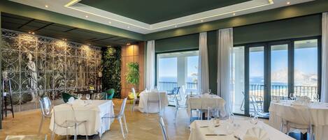 Restaurante com cadeiras e mesas de vidro transparente, um mural na parede e vista para o mar.