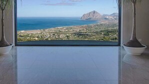 A room with a large window offering a view of a coastal landscape, two potted plants, and a tiled floor. - Venere di Erice Resort (Valderice)