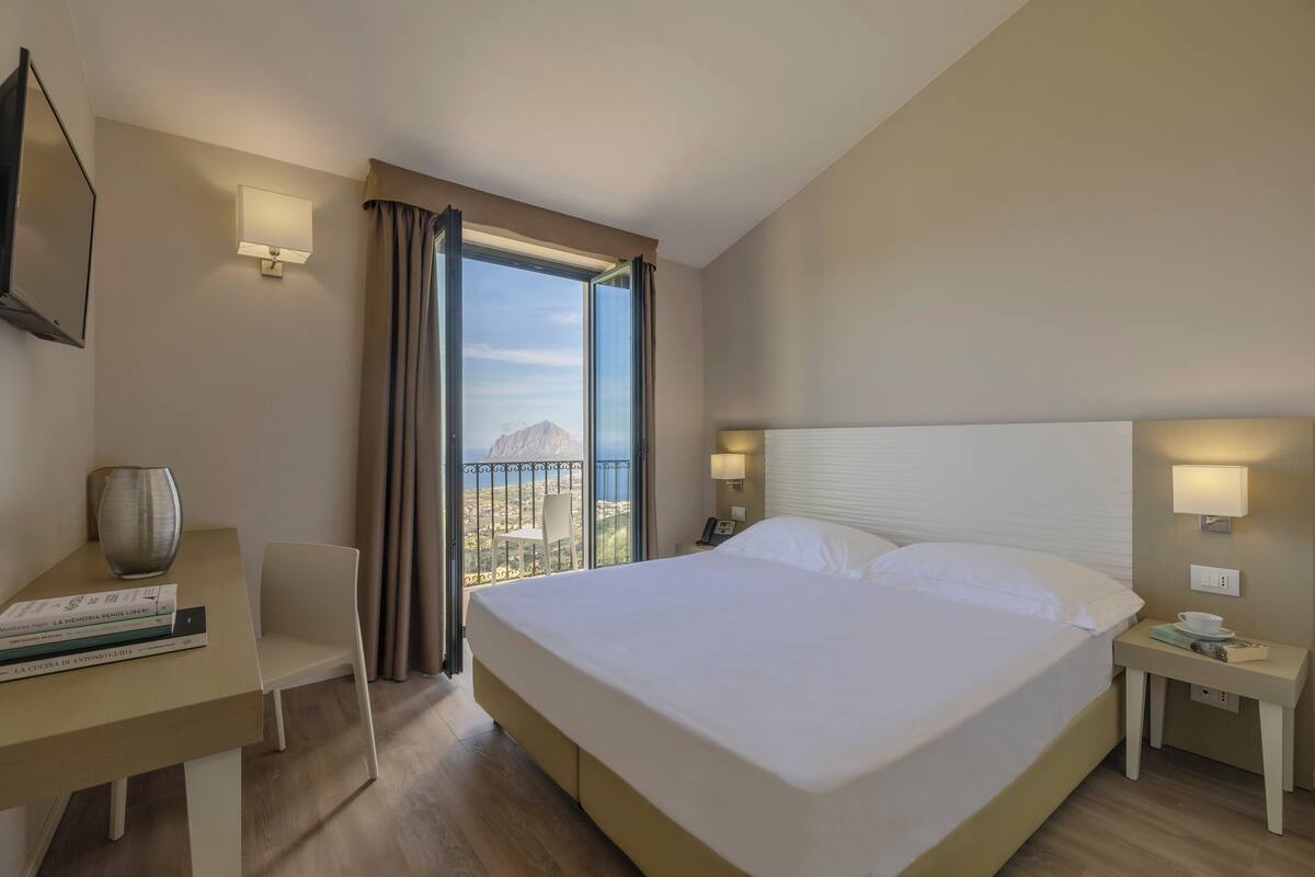 Habitación de hotel con una cama grande, un escritorio con una silla, una mesita y vistas a la ciudad a través de un ventanal amplio.