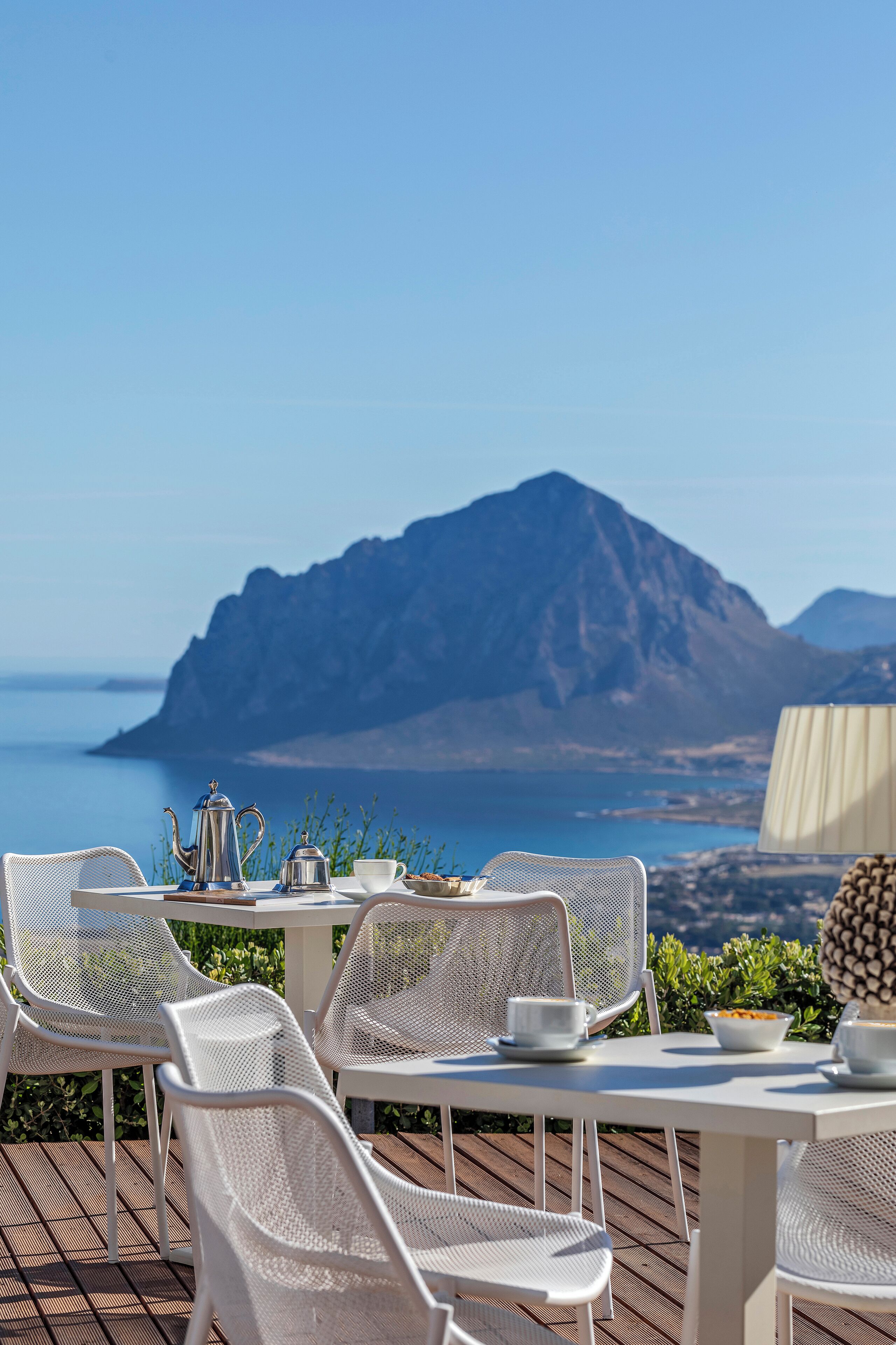 A table with white chairs and a table setting, including a teapot and cups, overlooking a scenic view of the sea and mountains.