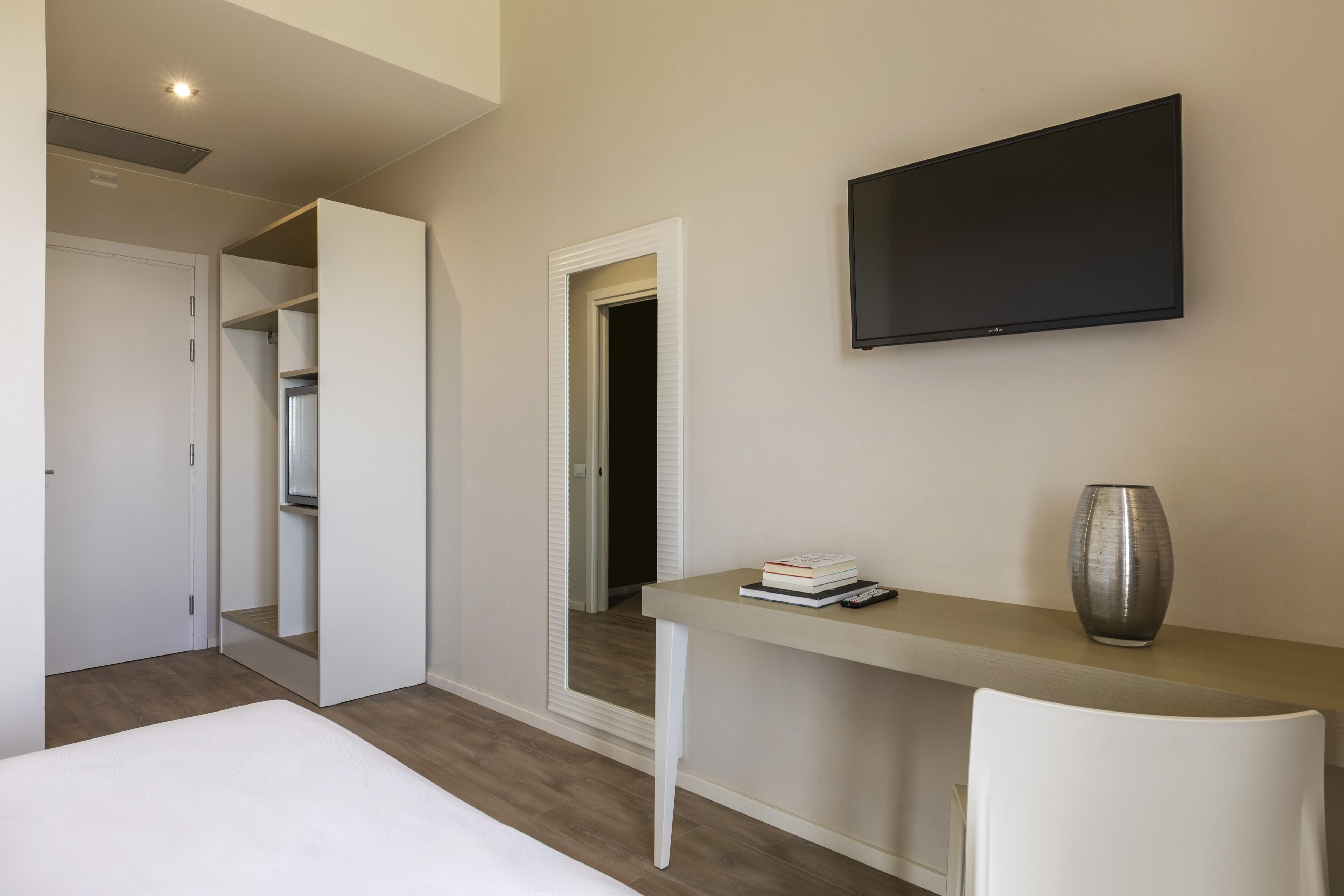 A modern hotel room with a bed, a desk with a vase, a flat-screen TV, and a walk-in closet.
