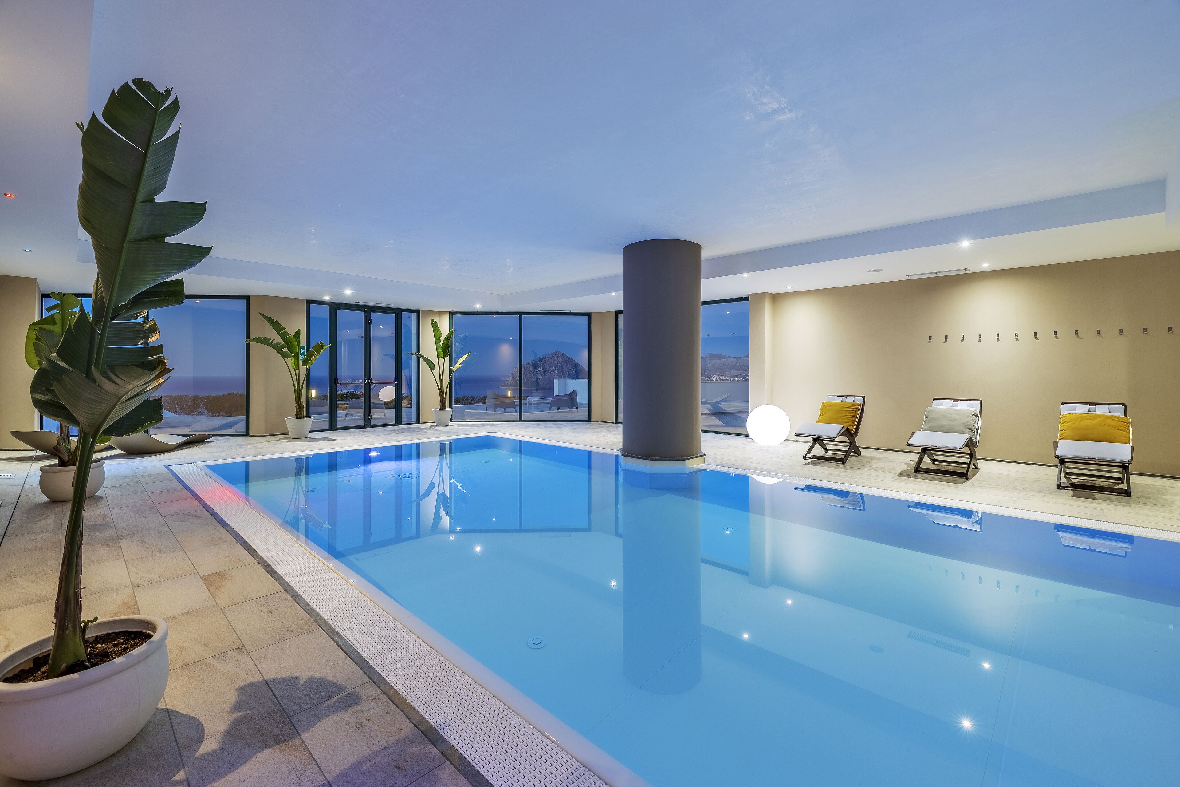 En indendørs swimmingpool med glasvægge, potteplanter og liggestole.