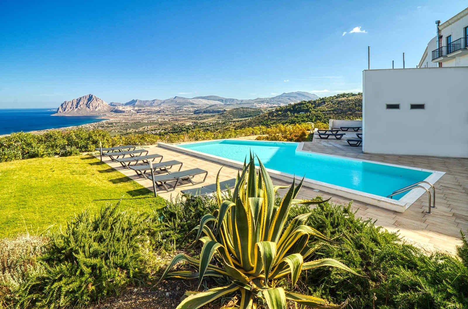 A swimming pool with lounge chairs, a view of the sea, and a mountainous landscape in the background.