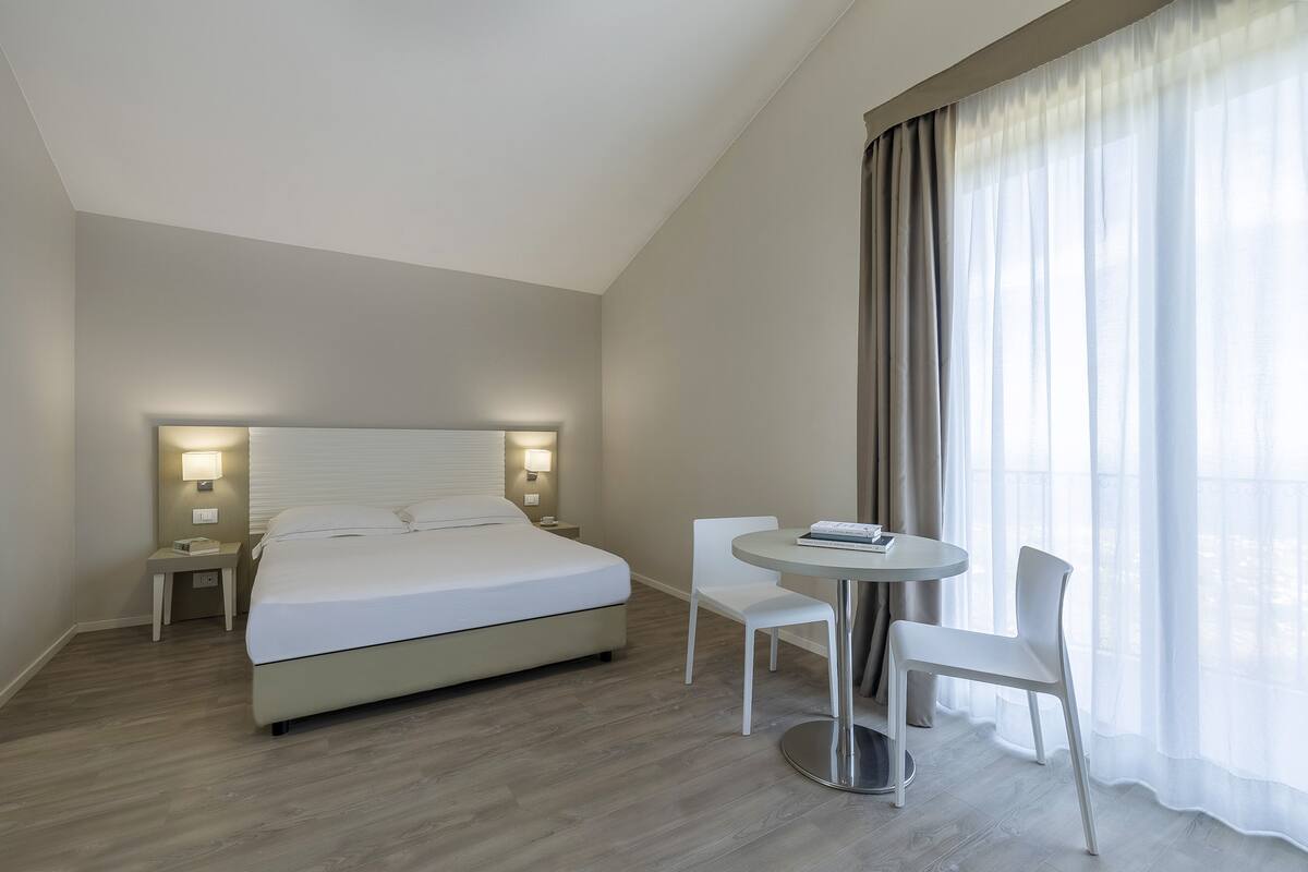 Habitación de hotel moderna con una cama grande, lámparas de noche, una mesa redonda con dos sillas y vistas al exterior a través de cortinas transl úcidas.