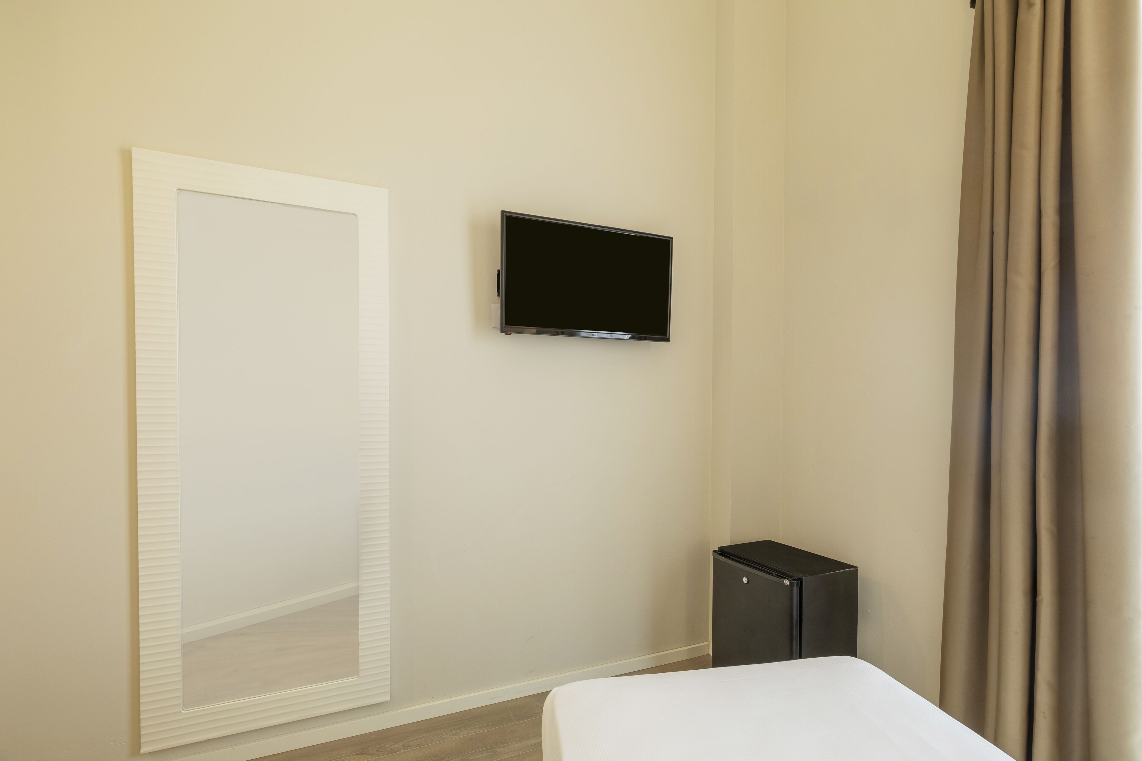 Una habitación de hotel con una cama, televisión fijada en la pared, un mueble pequeño y una ventana con cortinas.
