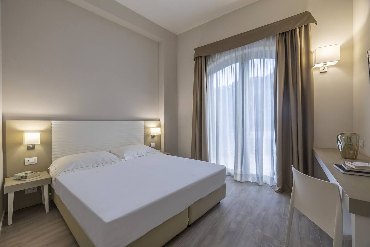 Habitación de hotel con una cama grande, lámparas de noche, un escritorio con silla y una ventana con cortinas.