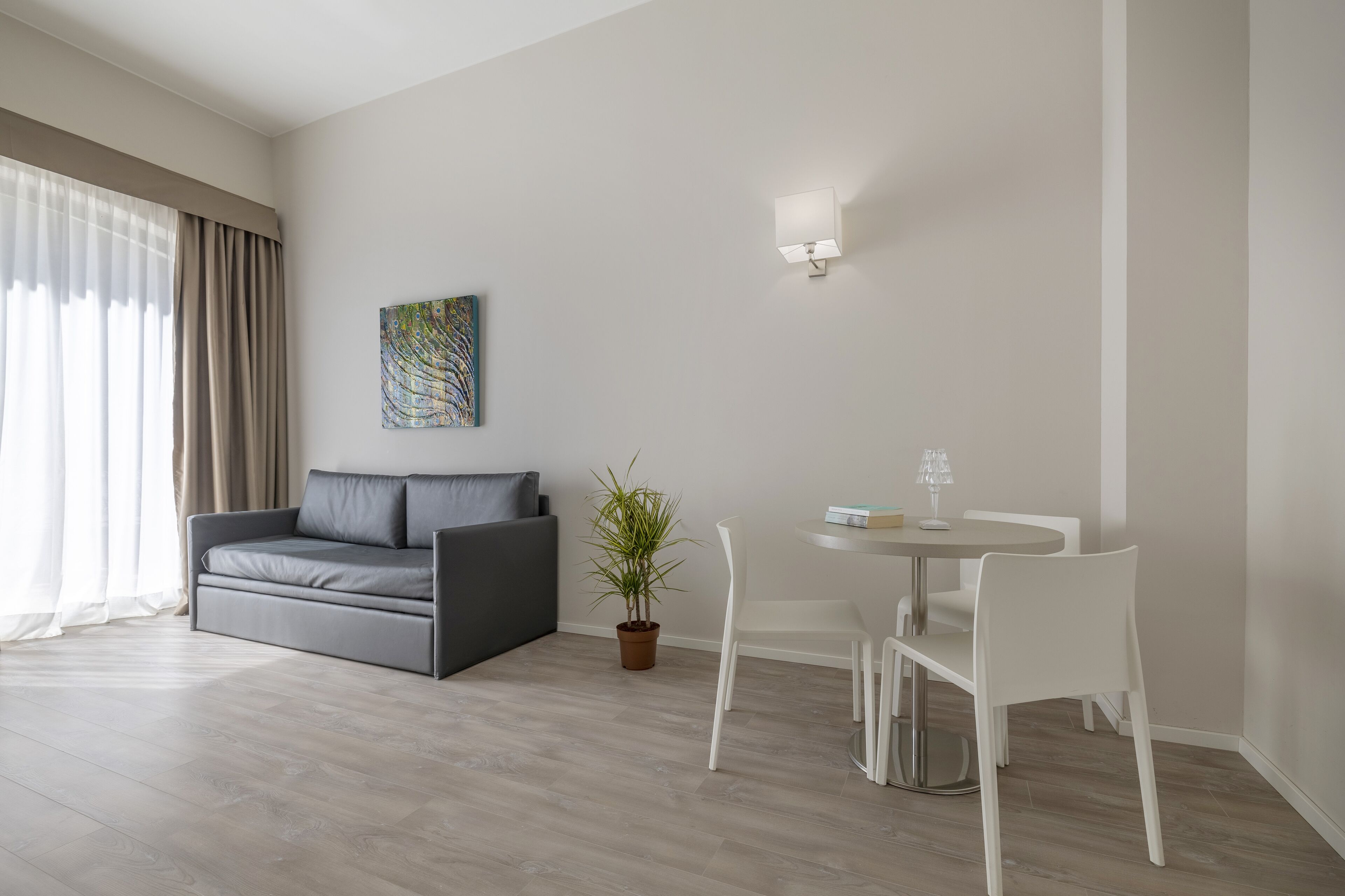 Ein modernes Wohnzimmer mit einem grauen Sofa, einem kleinen runden Tisch mit Stühlen, einer Topfpflanze und einer wandmontierten Lampe.