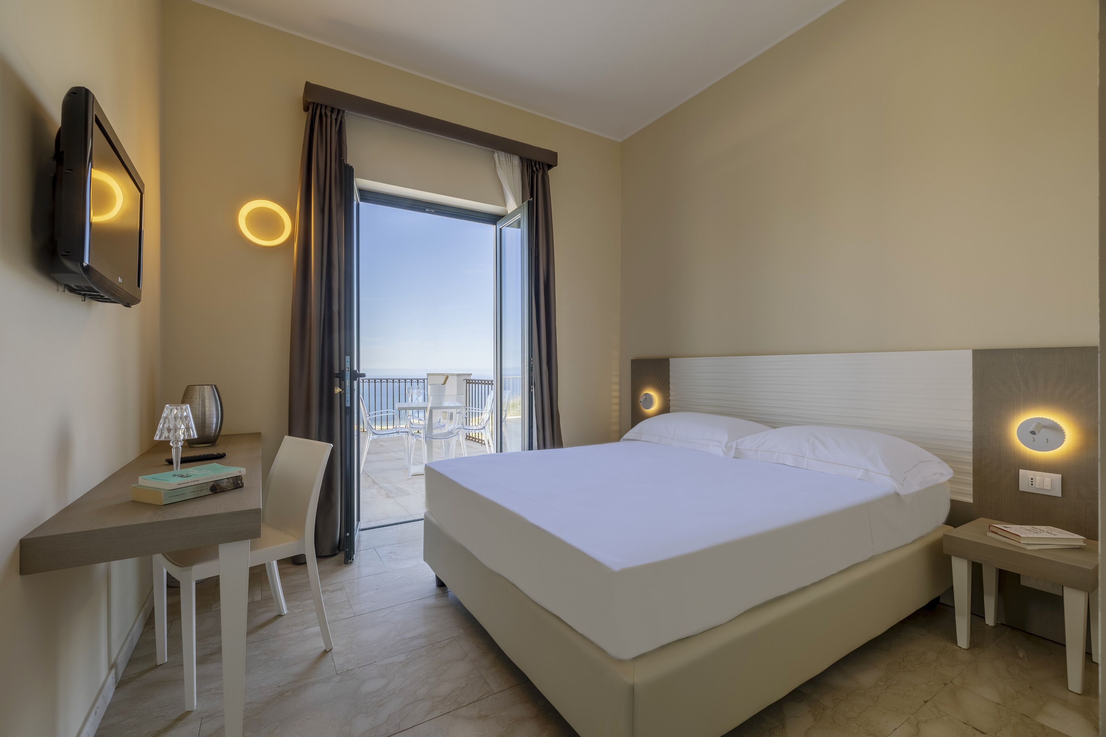 Quarto de hotel com cama, escrivaninha, cadeira e varanda com cortinas.