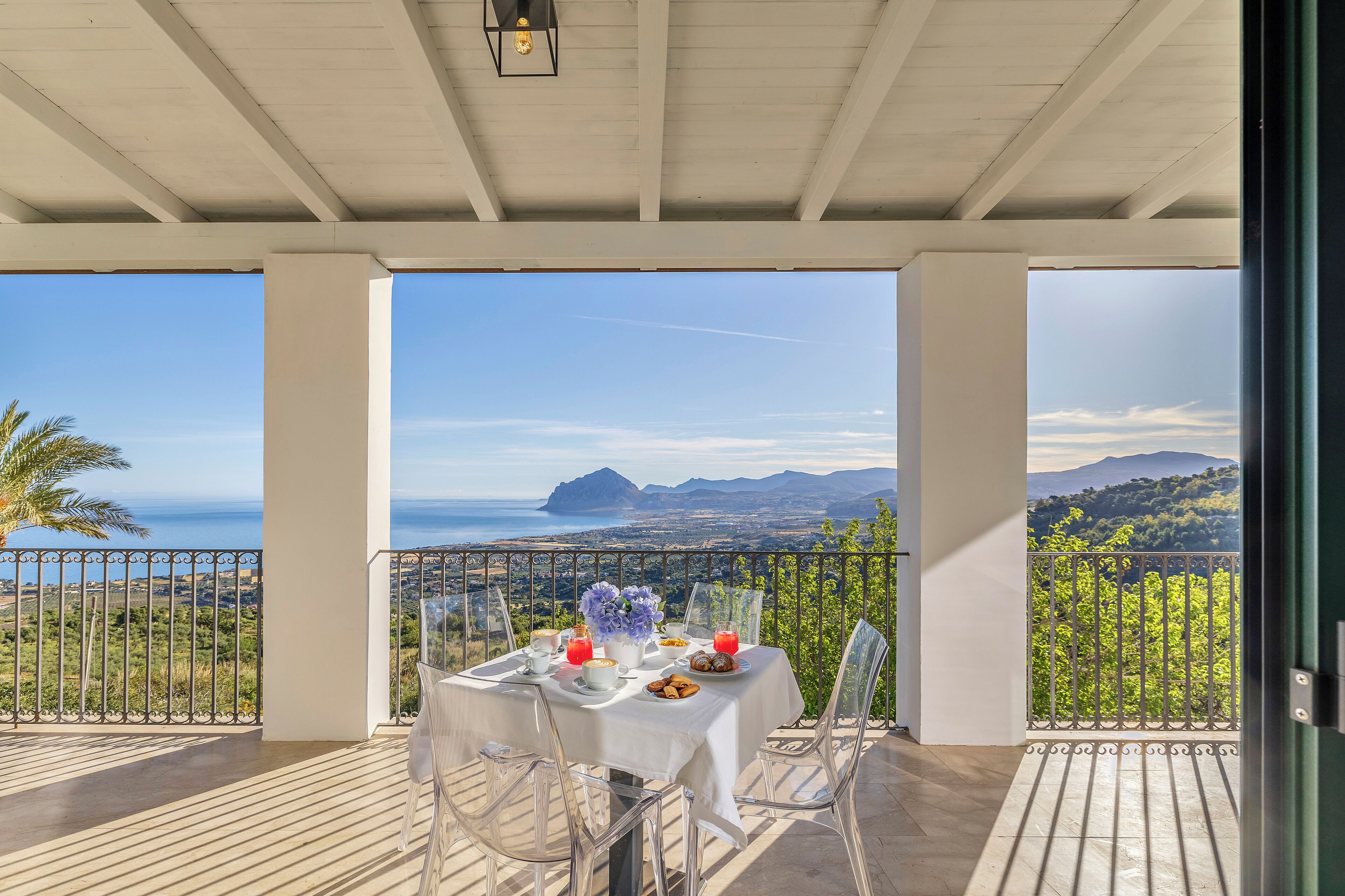 A balcony with a table set for breakfast, overlooking a scenic view of the sea and mountains.