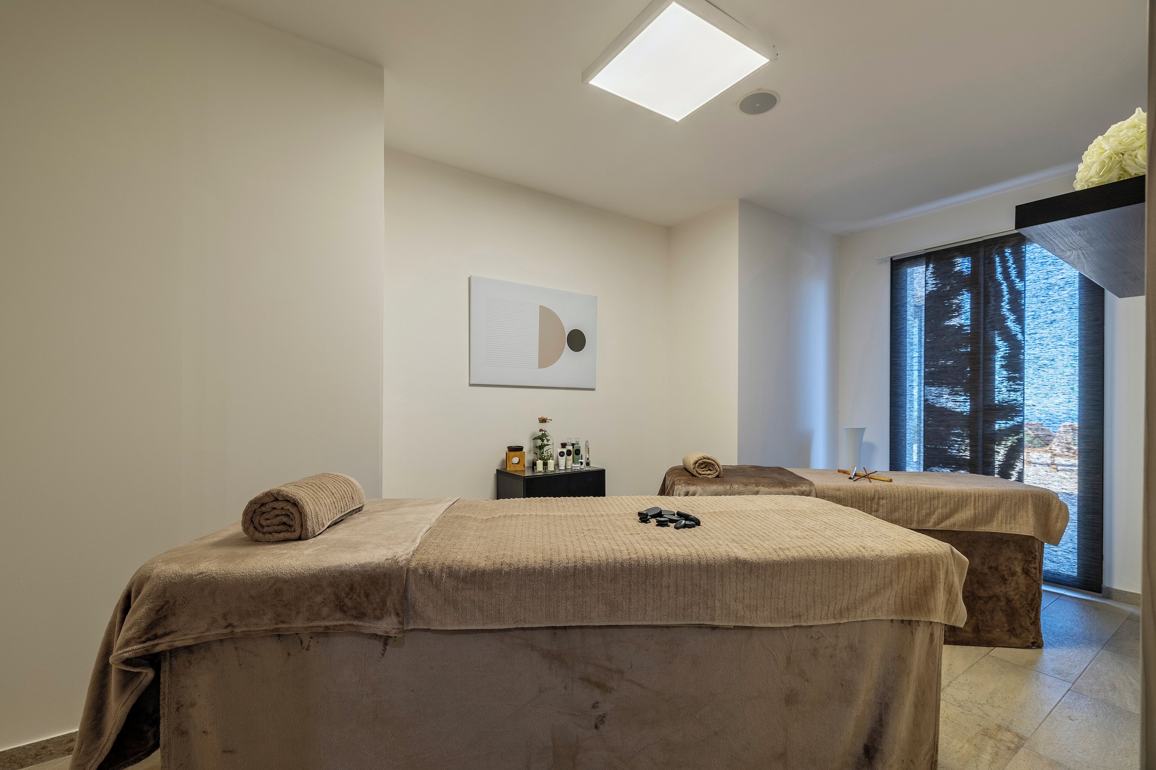 Une salle de spa équipée de deux lits, d’une petite table avec des articles de toilette et d’une grande fenêtre donnant sur un paysage agréable.