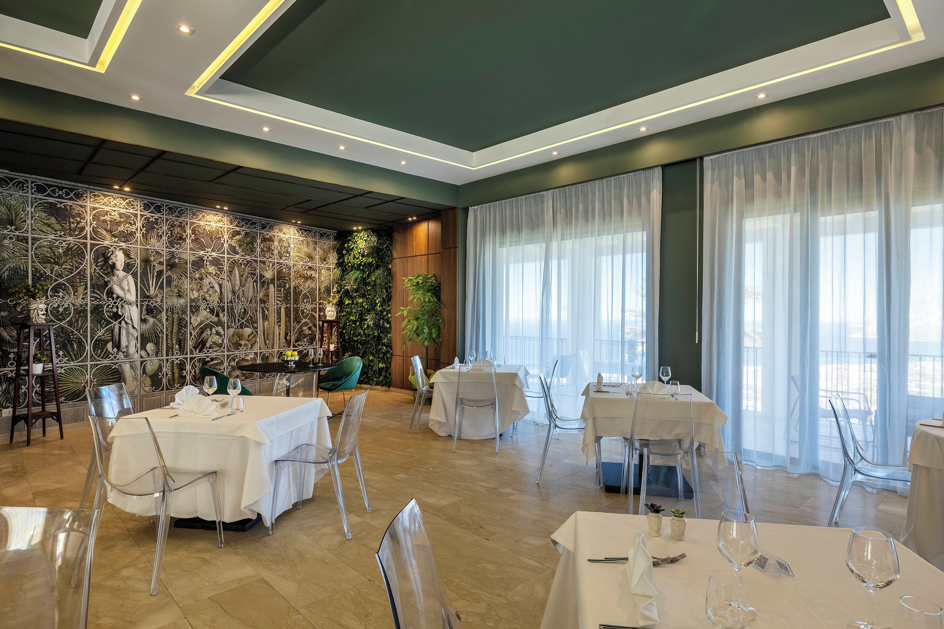 A restaurant with multiple tables, clear chairs, and a decorative wall with plants and a statue.