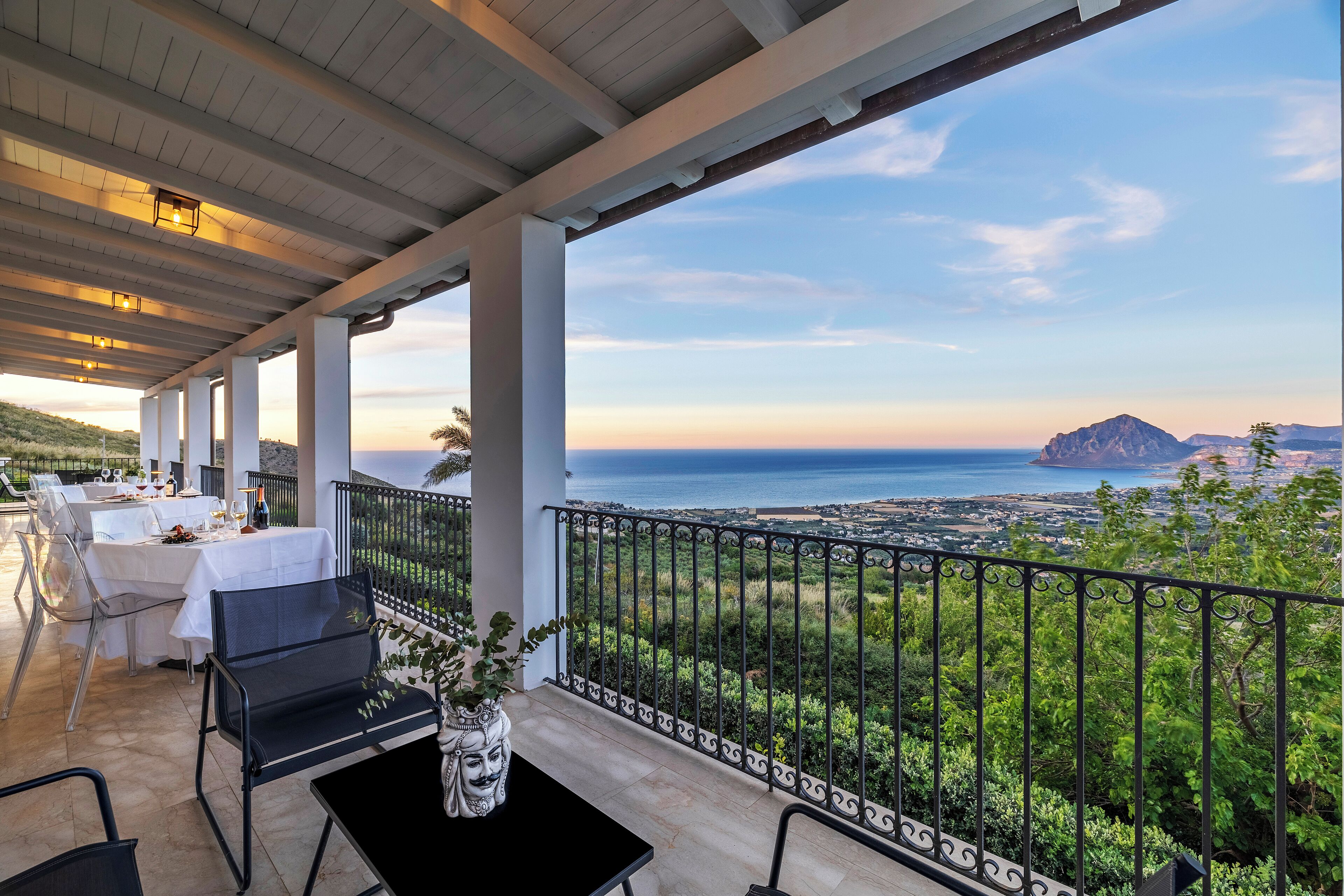 A covered outdoor dining area with tables and chairs, a potted plant, and a view of the ocean and distant land.