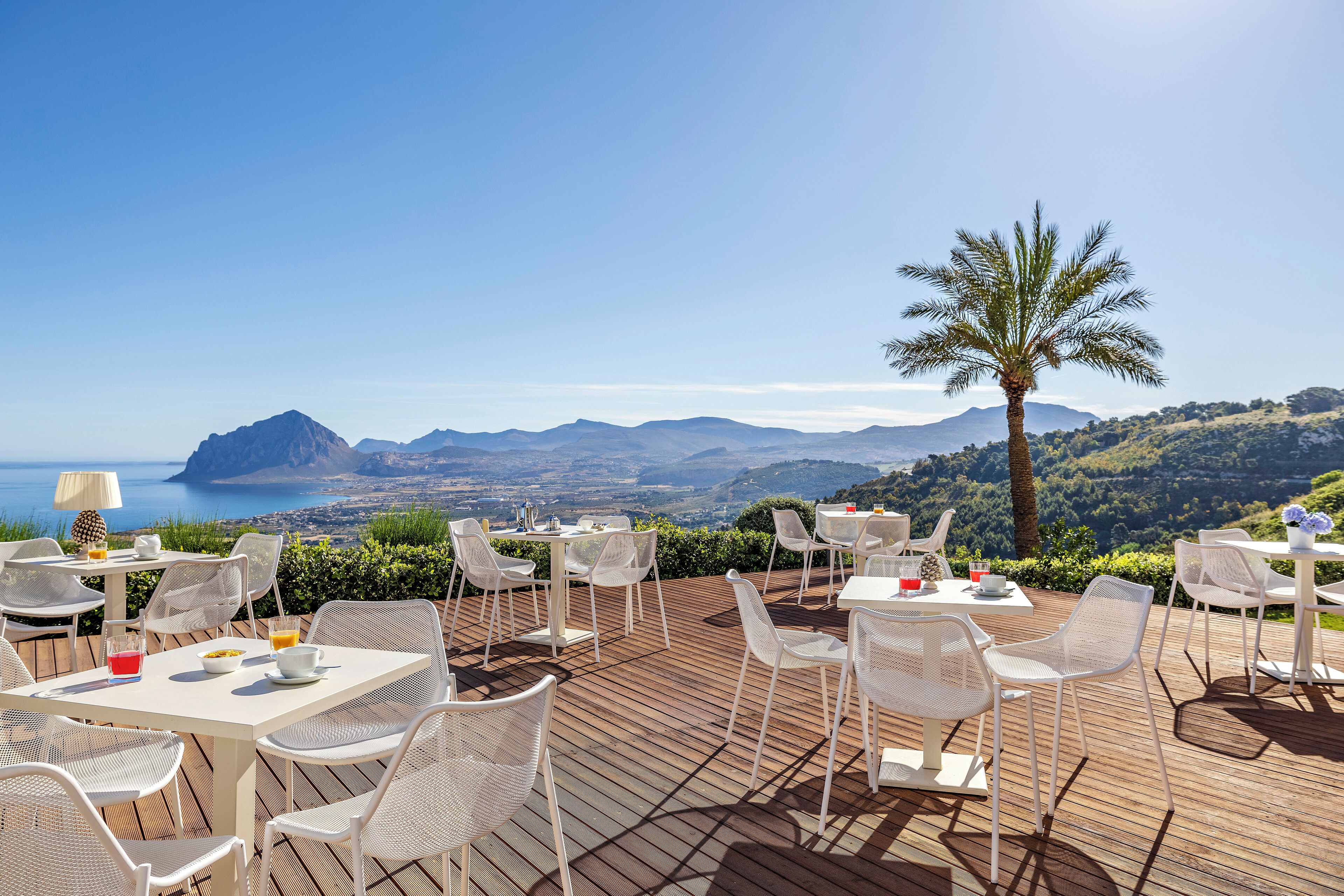 An outdoor dining area with white wicker furniture, tables, and chairs, overlooking a scenic landscape with mountains and the sea.