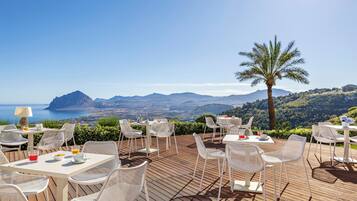 An outdoor dining area with white wicker furniture, tables, and chairs, overlooking a scenic landscape with mountains and the sea.