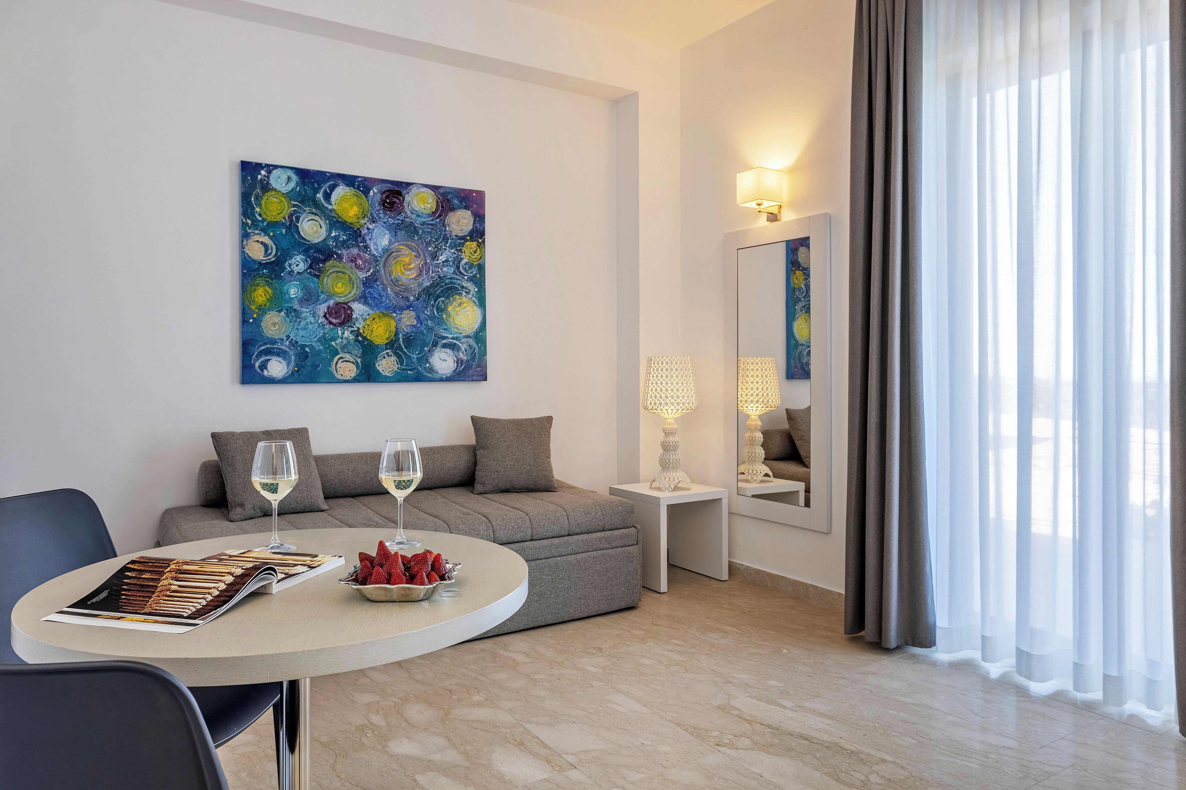Uma sala de estar moderna com um sofá, uma mesa de jantar com uma revista e morangos, um quadro na parede e um espelho refletindo o ambiente.
