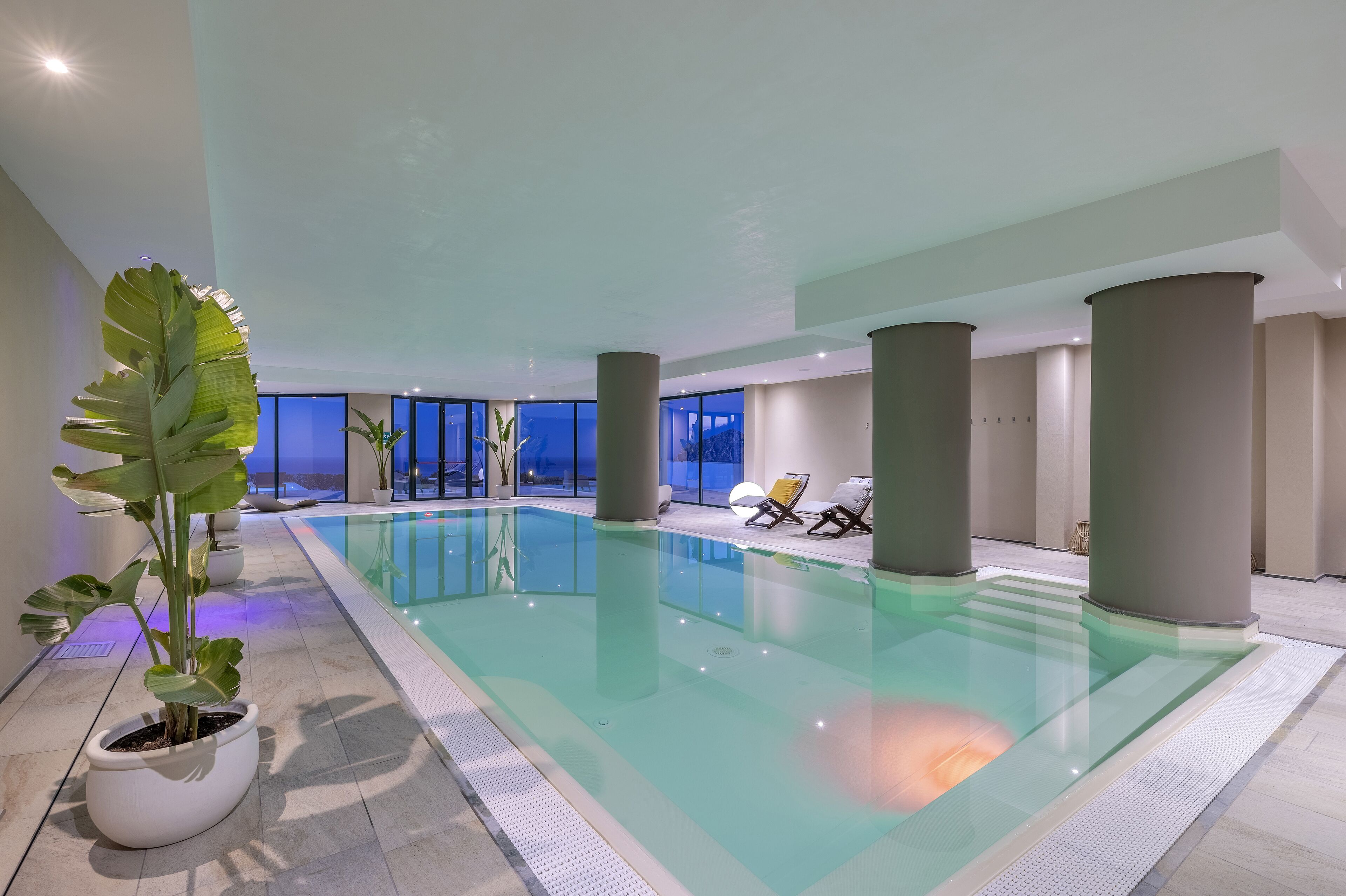 Una piscina interior con agua cristalina, rodeada de pilares grandes, plantas en macetas y sillas de descanso.