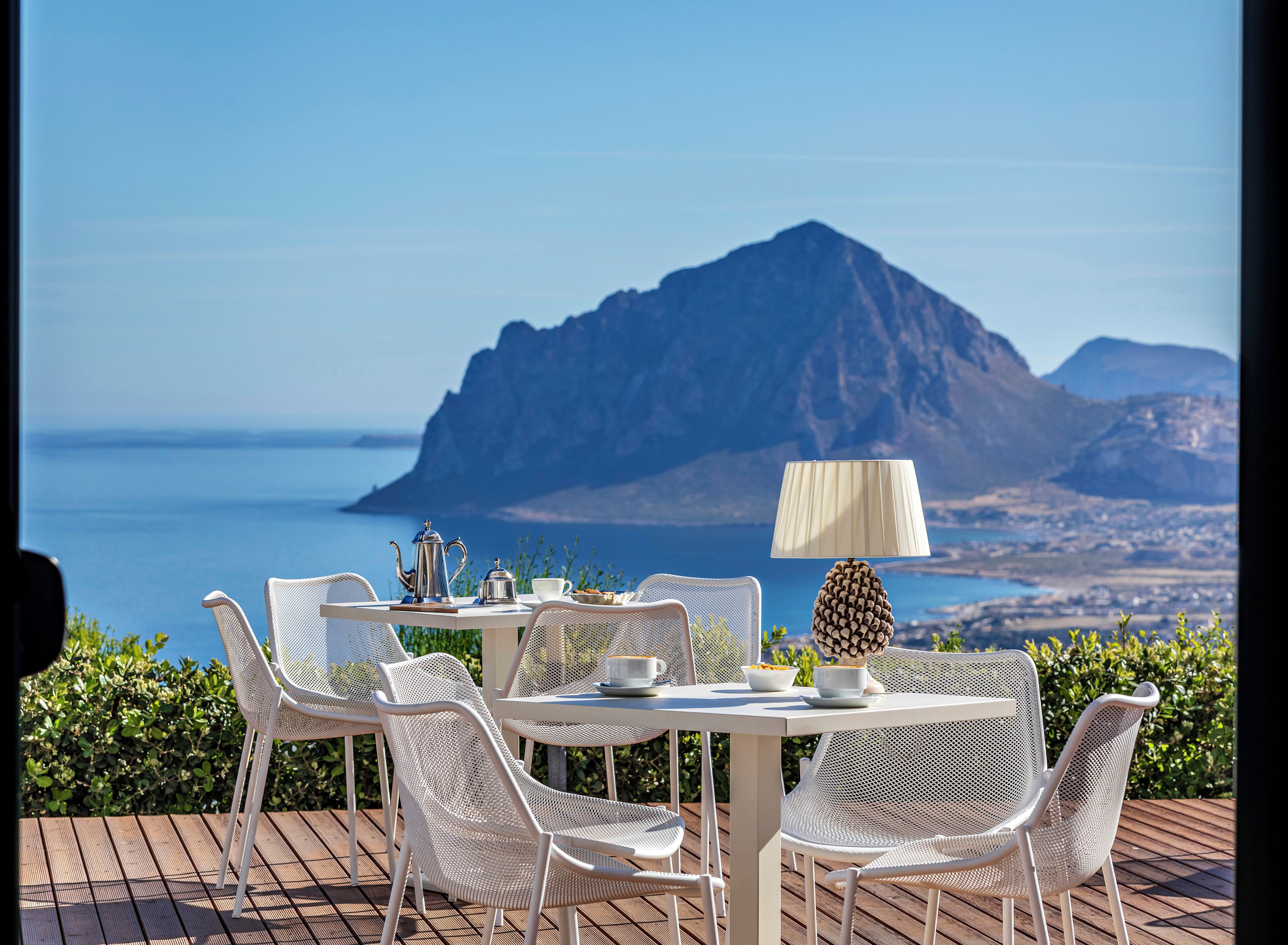 A table set for two with white chairs, a table lamp, and a view of the sea and mountains.