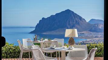 A table set for two with white chairs, a table lamp, and a view of the sea and mountains.