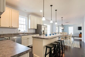 Uma cozinha moderna com armários brancos, bancada de granito, ilha central com banquetas, iluminação pendente e área de jantar com mesa e cadeiras.