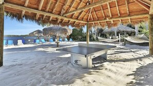 Una mesa de ping pong a la orilla de la playa, cubierta por un techo de paja, con sillas de playa y hamacas al fondo.
