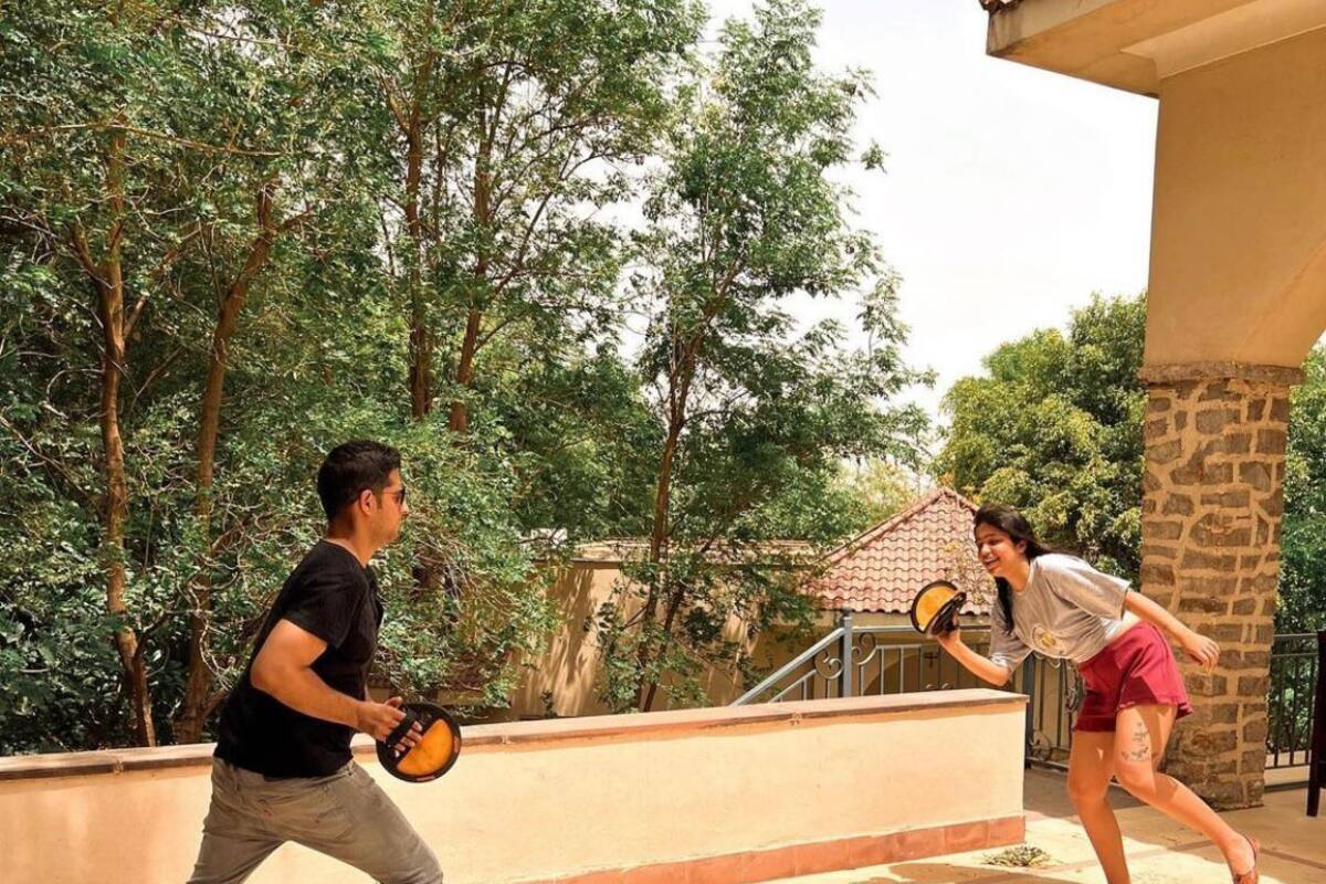 Two people playing badminton on a patio with a stone wall and trees in the background.