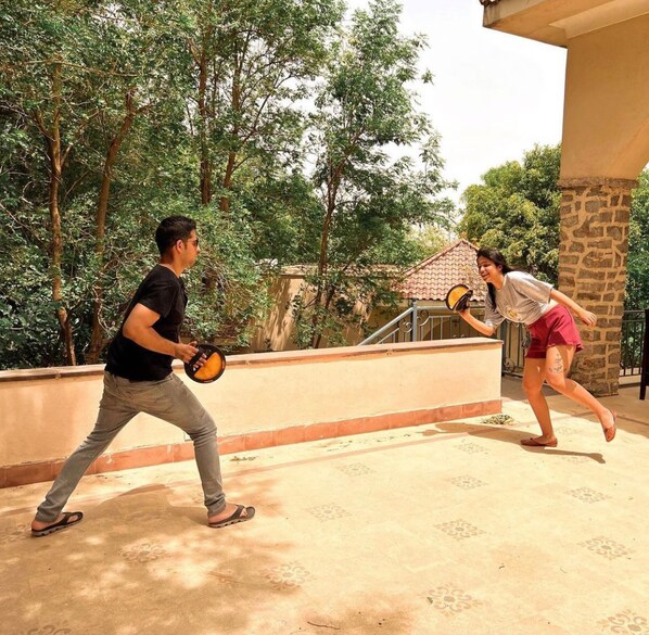 Due persone giocano a badminton su un patio con un muro di pietra e alberi sullo sfondo.