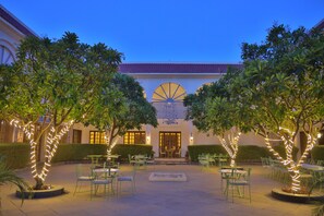 Un cortile esterno con tavoli e sedie, circondato da alberi e un edificio con un'ampia finestra ad arco.