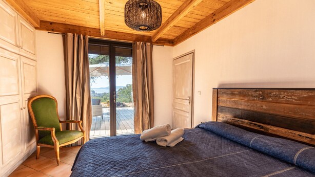 A bedroom with a wooden ceiling, a bed with a dark quilt, a green chair, and a view through a glass door. - Welcome to Filippi Furnished Bungalows! (PORTO-VECCHIO)