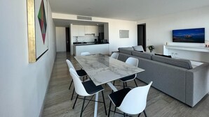 Ein modernes Wohnzimmer mit grauem Sofa, einem Esstisch mit weißen Stühlen, einer Küche im Hintergrund und einem an der Wand befestigten Fernseher.