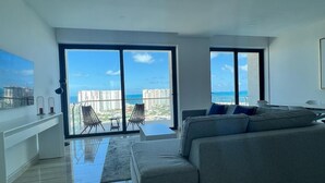Ein modernes Wohnzimmer mit einem großen grauen Sofa, einem Couchtisch und einem Sessel. Große Fenster bieten Blick auf die Stadt und die umliegenden Gebäude.
