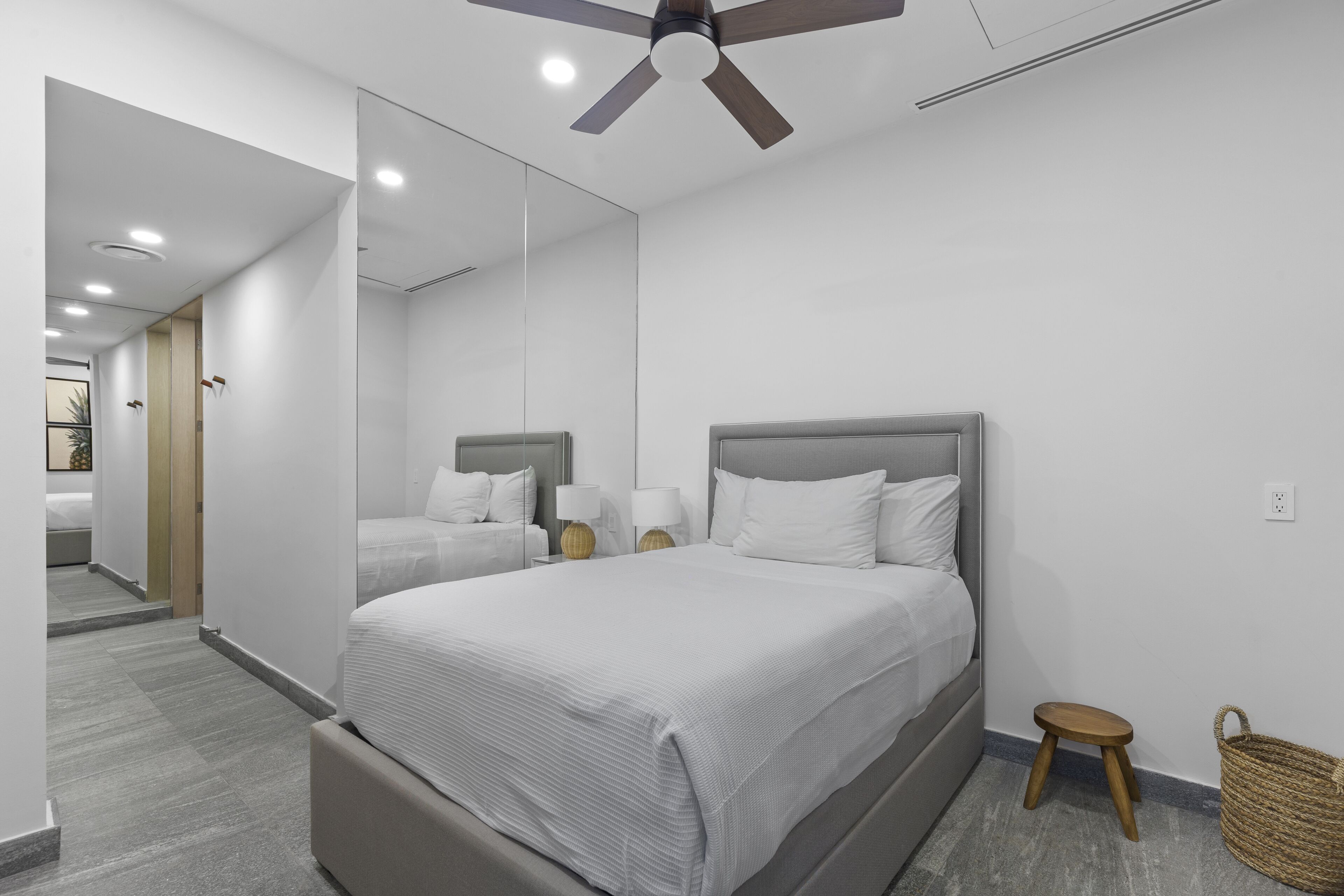 A modern hotel room with a large bed, a ceiling fan, a small stool, and a wicker basket.