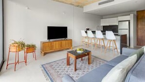 Een moderne woonkamer met een bank, een salontafel en een televisie. Er is een keukenhoek met een koelkast en een eethoek.